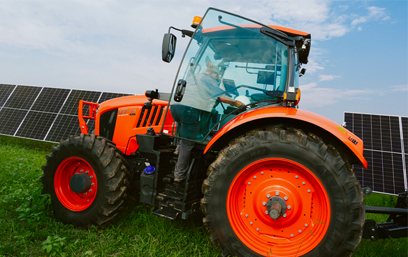 A tractor drives in front of rows of solar panels in a green field.